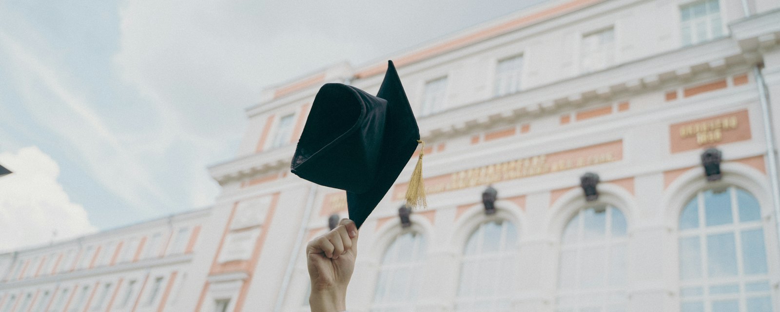 A graduate holding a mortarboard aloft outside a university building