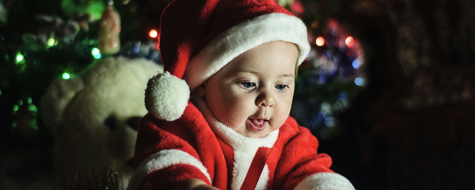 A baby in a red Santa outfit by the lights of a Christmas tree