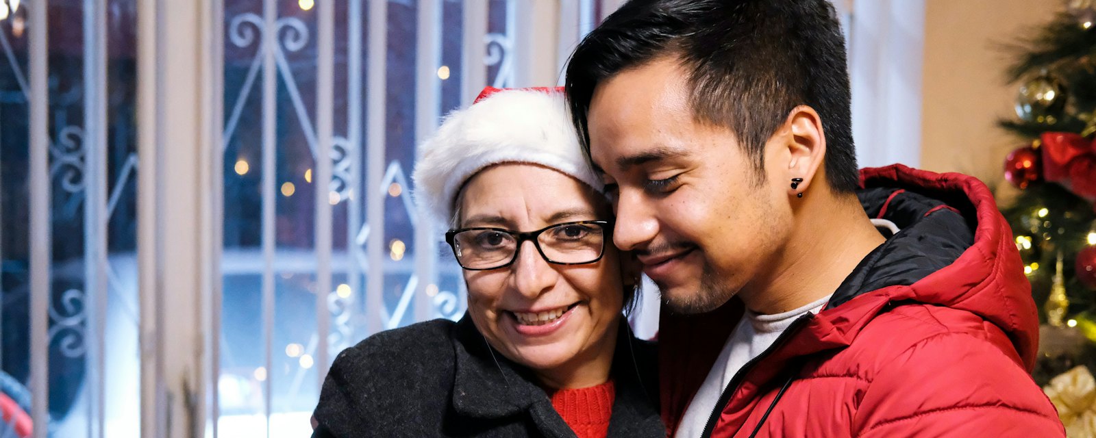 A grandmother in a Santa hat smiling with her grandson by a Christmas tree