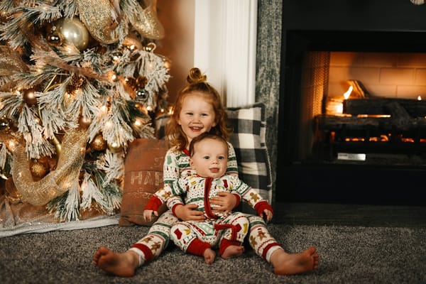 Two young children in matching Christmas pyjamas by a tree and fire